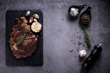 Dilimlenmiş biftek, 4 mevsimlik ızgara sarımsaklı biber, taş tabakta koyu gri arka plan, bıçak ve çatal siyahı, üst manzara