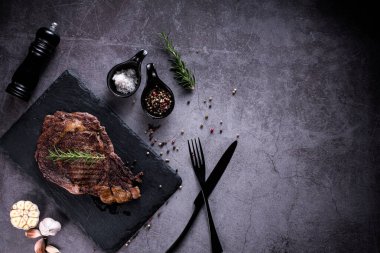 Dilimlenmiş biftek, 4 mevsimlik ızgara sarımsaklı biber, taş tabakta koyu gri arka plan, bıçak ve çatal siyahı, üst manzara