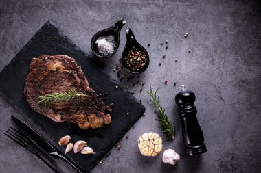 Dilimlenmiş biftek, 4 mevsimlik ızgara sarımsaklı biber, taş tabakta koyu gri arka plan, bıçak ve çatal siyahı, üst manzara
