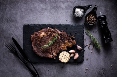 Dilimlenmiş biftek, 4 mevsimlik ızgara sarımsaklı biber, taş tabakta koyu gri arka plan, bıçak ve çatal siyahı, üst manzara