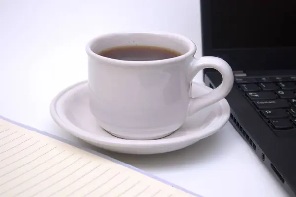 Bir fincan kahve, defter ve dizüstü bilgisayar beyaz masada.