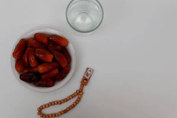 Beyaz bir zemin üzerinde bir tabak hurma ve tespih bulunan bir bardak maden suyu.