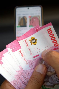 Adamın elinde 100.000 rupiah banknot var. Cep telefonundan online dükkanın geçmişi bulanık.