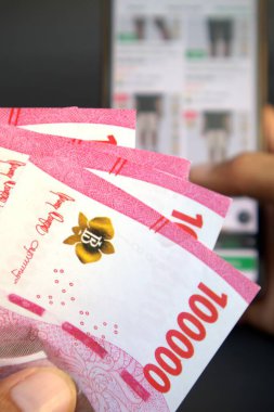 Adamın elinde 100.000 rupiah banknot var. Cep telefonundan online dükkanın geçmişi bulanık.