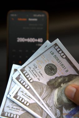 Adamın eli cep telefonunda bulanık arka plan hesap makinesiyle 100 Amerikan doları tutuyor.