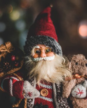 Neşeli bir Noel Baba figürü neşeli bir ortamda durur, parıldayan ışıklar, süsler ve güzel dekore edilmiş bir Noel ağacı ile çevrili, tatil sezonunun büyüsünü yakalar..