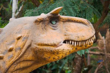Bir Carnotaurus düşük bitki örtüsünün arasında durur, kaslı yapısı vahşi araziyle harmanlanmış, gözleri çevreyi yırtıcı bir odak ile tarıyor..