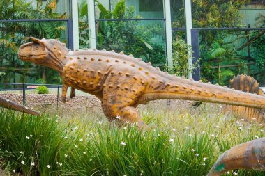 Bir Carnotaurus düşük bitki örtüsünün arasında durur, kaslı yapısı vahşi araziyle harmanlanmış, gözleri çevreyi yırtıcı bir odak ile tarıyor..