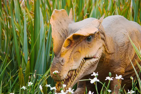 Bir Dilophosaurus, orta büyüklükteki bitkiler ve dağınık kır çiçekleriyle çevrili yemyeşil çimlerin arasında duruyor. Tarih öncesi ortama uyum sağlıyorlar..