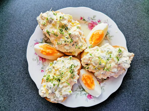 stock image Sandwich bread with egg spread with eggs. White plate with floral pattern. Three slices of bread with egg spread. Sprinkled with watercress. Served with three pieces of egg. Dark kitchen.