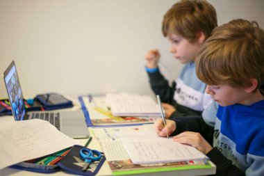 İki çocuk bir masada dizüstü bilgisayar ve kitaplarla ders çalışıyor.