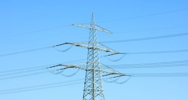 Açık bir gökyüzü önündeki devasa yüksek voltajlı elektrik direği, modern altyapılar - Münih, Almanya, Avrupa - için istikrarlı enerji şebekelerinin kritik rolünü sembolize ediyor.