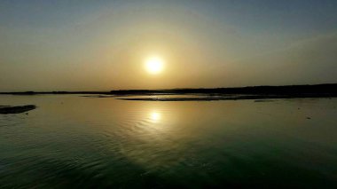 Güneş merkezli bir su kütlesinin üzerinde sakin bir günbatımı. Yüzeyde yansıması, sakin ve simetrik bir sahne yaratıyor.