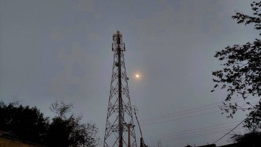 Antenleri gri gökyüzüne ve görünür aya karşı duran uzun bir baz istasyonu siluetli ağaçlar ve elektrik hatları tarafından çerçevelenmiş sakin bir akşam sahnesi teknolojisi ve sakin doğayla harmanlanmış..