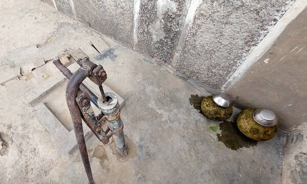 An old hand water pump stands in the corner of a village home's concrete structure, showing rust and wear, with two clay pots beside it for collecting water. The pump appears weathered and possibly not fully functional