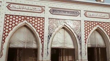 Beyaz cepheli güzel tasarlanmış bir cami, beş kemerli giriş ve kareli desenler, çiçek motifleri ve Arapça kaligrafiyle süslenmiş üç minare. Kırmızı ve yeşil dua paspasları öndeki açık alanı kaplıyor.