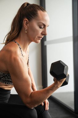 Spor salonunda güç antrenmanının ortasında bir fitness kadını, bir dambılı kaldırırken odaklanma ve kararlılığını gösteriyor..