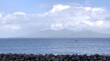 Deniz anakayası ve karanlık kumla güzel bir sahil manzarası. Arka planda dağ manzarası, bulutlu ve güneşli gökyüzü. Koyu tortu deniz kıyıları ve berrak su..