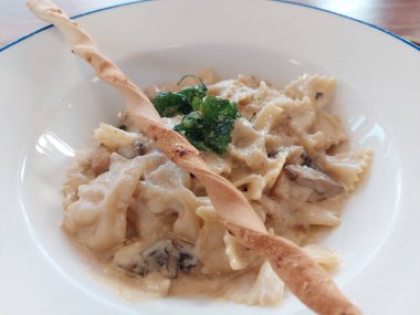 Spagetti Tavuklu Mantarlı Carbonara beyaz bir tabakta servis edilir. Tahta bir masada bir fincan çay ile..