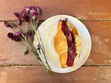 Peynir ve pastırmalı kruvasan, beyaz bir tabak üzerinde servis ediliyor, papatya çiçeği de ahşap, eski bir şarap masasının üstünde. Klasik estetik gıda fotoğrafçılığı.