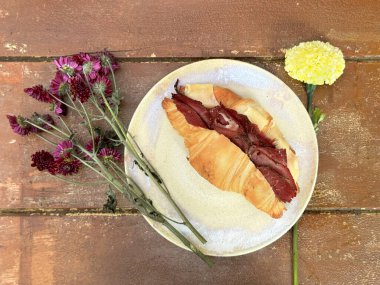 Cheese and bacon baked croissant served on a white saucer plate, with daisy flower on top of wooden vintage rustic table. Vintage aesthetic food photography. clipart