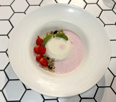 Çilekli panna Cotta, pembe krema, yulaf ezmesi ve tahıl gevreği. Beyaz bir altıgen mozaik masada beyaz bir tabakta servis edilmiş..
