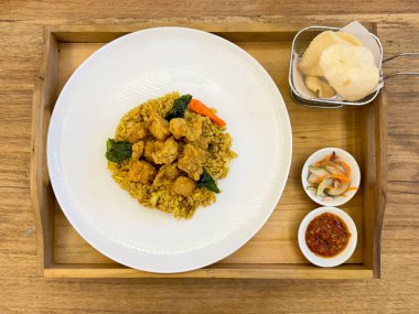 Otantik Endonezya otları ve baharatlı pirinçle tavuklu rendang. Tahta bir tepside, ahşap bir masada karides kraker, marine edilmiş salatalık ve acı sos sosuyla servis edilir..