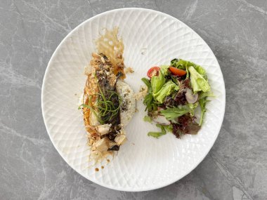 Baharatlı pilav ve Sezar salatası ile ızgara balık uskumrusu. Beyaz yuvarlak bir tabakta, gri mermer bir masada servis edilir..