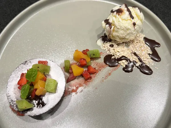 Vanilyalı dondurma kepçeli çikolatalı kek, granola, meyve ve pudra şekerli lav keki. Siyah bir masada gri bir tabakta servis edildi..