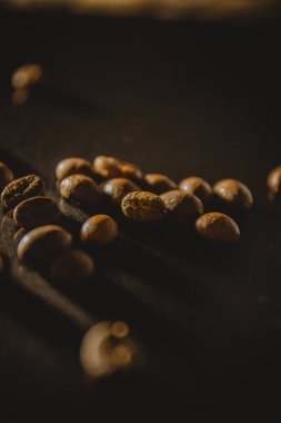 Fotoğraf koyu bir arka planda birkaç kahve çekirdeğini gösteriyor. Kahve çekirdeklerinden biri ön planda yer alır, açıkça onun dokusuna ve detaylarına dikkat çekerken, diğer fasulyeler ise bir aydınlatmaya yerleştirilir.
