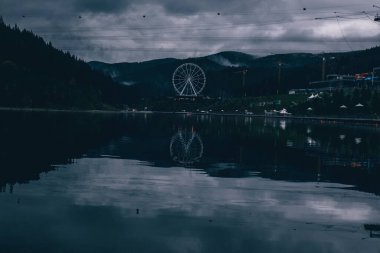 Yağmurlu havada dağların ve göllerin arka planına karşı büyük bir gözlem çarkı. Sakin sularda yansıma, gecenin sessiz renkleri, mistik atmosfer.