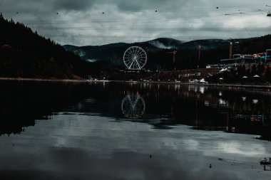 Yağmurlu havada dağların ve göllerin arka planına karşı büyük bir gözlem çarkı. Sakin sularda yansıma, gecenin sessiz renkleri, mistik atmosfer.