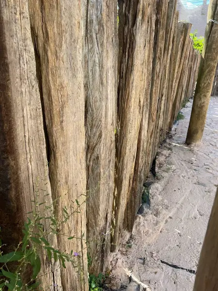 Köy ahşap bir çitin yakın görüntüsü, aşınmış odunlardan yapılmış, tabanında yeşillik bir bakış ve yanında kumlu bir yol, doğal ve sakin bir açık hava ortamını çağrıştırıyor..