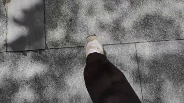 Siyah pantolonun altında spor ayakkabılarla POV üst görüş bacağı güneş ışığı altında ağaçların gölgesiyle lekeli gri seramik fayanslara basıyor. Yemekten sonra yürüyüp yemeği hazmetmek ve zamanın tadını çıkarmak..