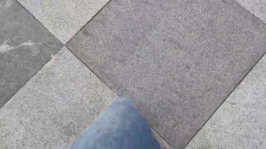 Mavi kot pantolonun altındaki siyah spor ayakkabılı POV üst görüş bacağı parkın altındaki eşkenar dörtgen seramik fayanslara basıyor. Yürüyüş ve egzersiz konsepti. İlk adım atma görüntüsü.