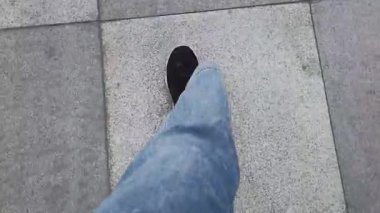Mavi kot pantolonun altındaki siyah spor ayakkabılı POV üst görüş bacağı parkın altındaki kare seramik fayanslara basıyor. Yürüyüş ve egzersiz konsepti. İlk adım atma görüntüsü.
