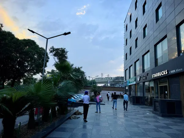 Gurugram, Hindistan - 5 Ağustos 2024: Starbucks yakınlarındaki akşam sokak sahnesi, insanlar arka planda metro ile ağaçlı kaldırımda geziniyorlar. Güzel mavi gökyüzü şehir ortamını güçlendirir