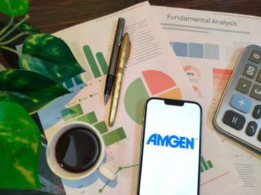 New York, ABD - 15 Aralık 2024: Finansal içgörü için akıllı telefonunda AMGEN logosu bulunan grafikler, grafikler, hesap makinesi ve kahvenin yer aldığı AMGEN borsa analizi üzerine seçici odaklanma
