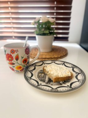 Renkli çiçekli bir fincan kahve ve süzme peynirli tostlu bir tabağın yer aldığı rahat bir kahvaltı. Arka planda çiçek saksısı olan huzurlu bir sabah havası..