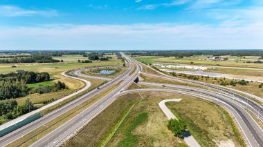 Otoyol kesişiminin doğal, bastırılmış renklerle çekilmiş havadan çekilmiş fotoğrafı. Sahne altyapı ve doğanın karışımını vurguluyor, sakin bir banliyö manzarası boyunca uzanan yollar..