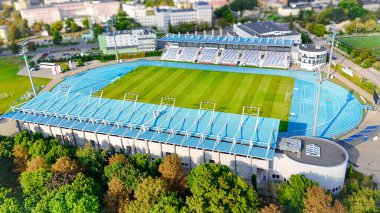 Yeşillik ve şehir manzarasıyla çevrili bir futbol sahası ve koşu pistinin yer aldığı modern bir spor stadyumunun yüksek kaliteli hava manzarası. Spor, mimari ve hava fotoğrafçılığı temaları için ideal.