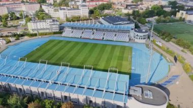Görüntü, havadan çekilmiş modern bir spor stadyumunu gösteriyor. Yeşil futbol sahası çarpıcı mavi bir atletizm pisti ile çevrilidir. Mavi aksanlı tribünler stadyumun her iki tarafından da görülebiliyor..
