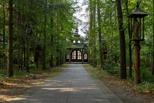 stock image A serene statue of a saint is sheltered under a small roof at the entrance of a shrine. The shrine is surrounded by tall, dense trees, and a brick wall complements the wooden structure. The peaceful forest setting enhances the spiritual ambiance 