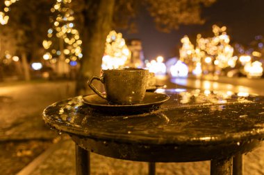 Close-up of a cup of coffee and a slice of cake on a wet table at night. Festive golden bokeh lights create a warm atmosphere. Ideal for holiday, winter, or cozy night-themed projects. clipart