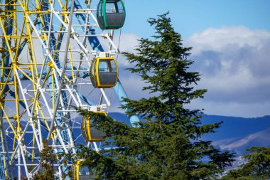 Mtatsminda Park, Tiflis, Georgia 'daki dönme dolabın canlı bir görüntüsü panoramik şehir manzarası sunuyor. Görüntü, renkli tekerleği parkların arka planına, yemyeşil alana ve şehrin siluetine yansıtıyor. Popüler bir turist sergileniyor
