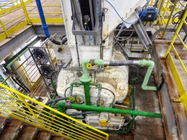 Görüntü, çeşitli borular ve vanalarla bağlı bir pompa ya da kompresöre benzeyen endüstriyel bir mekanizmayı gösteriyor. Ekipmanlar ağırlıklı olarak metalik ve rendelenmiş bir zemine kurulmuş.. 