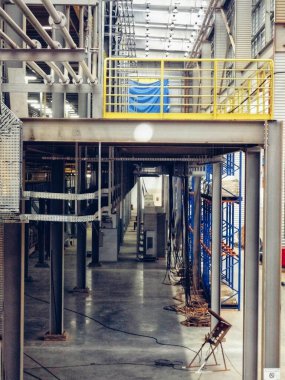 Görüntü bir sanayi ortamını gösteriyor, muhtemelen bir fabrika ya da bir depo. Metal yapılar, tavan boyunca uzanan borular ve makine ya da üretim hattının parçaları gibi çeşitli elementler var..