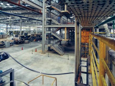Görüntü bir sanayi ortamını gösteriyor, muhtemelen bir fabrika ya da bir depo. Uzaya dağılmış çeşitli ağır makineler ve ekipmanlar var. Tavan yüksek metal kirişler ve floresan ışıklarıyla. 