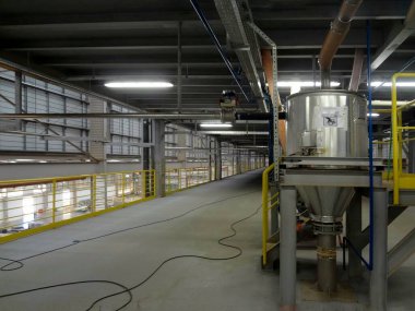 Görüntü bir sanayi ortamını gösteriyor, muhtemelen bir fabrika ya da işleme tesisi. Sarı parmaklıklı metal yapılar var, muhtemelen güvenlik için. Resmin sağ tarafında konik tabanlı büyük silindirik bir kap belirgindir..