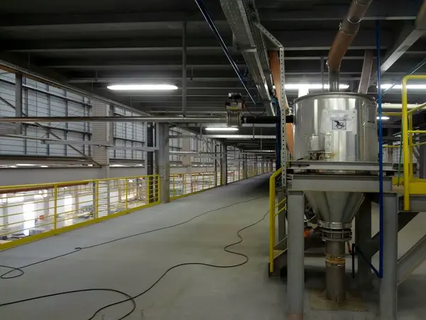 Görüntü bir sanayi ortamını gösteriyor, muhtemelen bir fabrika ya da işleme tesisi. Sarı parmaklıklı metal yapılar var, muhtemelen güvenlik için. Resmin sağ tarafında konik tabanlı büyük silindirik bir kap belirgindir..